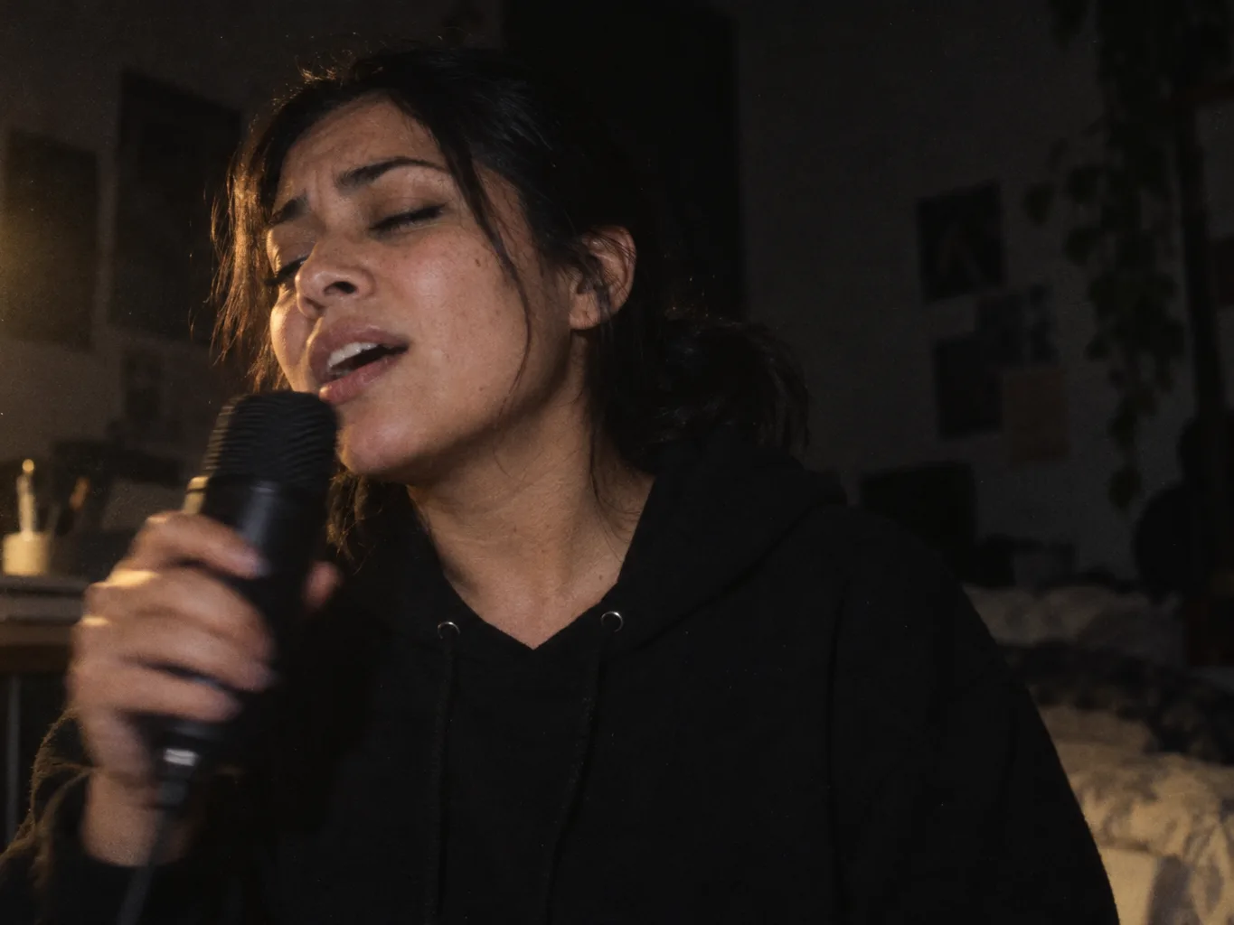 A woman in a hoodie singing into a microphone in a dim bedroom, eyes half closed
